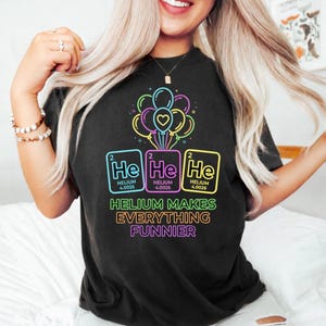 Könnte beinhalten: Ein schwarzes T-Shirt mit einer Grafik aus bunten Luftballons im Neon-Stil über drei Periodensystem-Quadraten für Helium, beschriftet mit He, Helium und 4.0026. Unter der Grafik steht der Text: HELIUM MAKES EVERYTHING FUNNIER in Neonbuchstaben.
