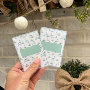 May include: Two rectangular cards featuring a repeating pattern of light blue dinosaurs and green tufts of grass on a white background. Each card has a solid, light green horizontal rectangular label centered on the front for writing text.
