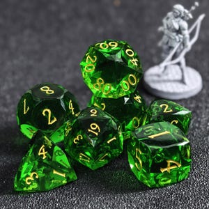 Puede incluir: Un conjunto de dados poliédricos de color verde esmeralda translúcido con números dorados. La colección incluye varias formas como d20, d12, d10, d8, d6 y d4, todas con bordes afilados y un acabado pulido, exhibidas junto a una pequeña figura gris.