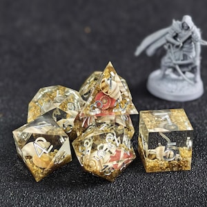 May include: A set of polyhedral dice made of clear resin, each containing embedded miniature seashells, sand, and small red accents. The dice feature silver-colored numbers and are arranged in a cluster, with a small gray figurine visible nearby.