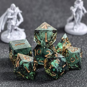 May include: A set of polyhedral dice made of dark green resin with swirling white smoke-like patterns and embedded gold-colored flakes. Each die features recessed gold-colored numbers. The set includes various geometric shapes used for tabletop gaming.