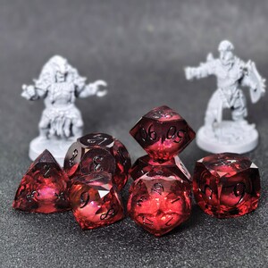 Puede incluir: Un conjunto de dados poliédricos de color rojo translúcido con numeración negra, bordes afilados y superficie facetada. La colección incluye varias formas como d20, d12, d10, d8, d6 y d4, dispuestas en un grupo con dos figuras en miniatura.
