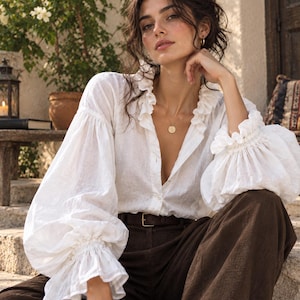 May include: A white long-sleeved button-down shirt featuring a ruffled collar and voluminous sleeves with ruffled cuffs, paired with dark brown trousers and a simple brown leather belt with a gold-toned buckle, accessorised with a gold pendant necklace.