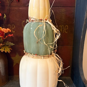 May include: A vertical stack of three decorative pumpkins in cream, sage green, and off-white. The pumpkins are adorned with thin brown vines, small red berries, and natural raffia ribbon tied around the middle and top sections of the stack.