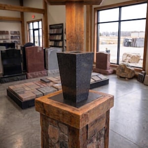 May include: A dark gray granite vase with a tapered square shape sits atop a rustic wooden pedestal. The pedestal features a stone-clad base and a thick wooden top. A small light fixture is recessed into the wooden structure above the vase.
