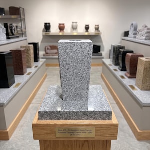 May include: A grey tapered granite vase with a speckled texture, resting on a matching square granite base. A brass plaque on the wooden pedestal reads: Item 459 - Midwestern Stone Supply: Premium Tapered Granite Vase - Grey (4"x5"x9").