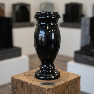 May include: A polished black granite vase with a smooth, rounded body and a flared rim, featuring a reflective surface. The vase is positioned on a rustic wooden block with a small metal plaque that reads MIDWESTERN STONE SUPPLY.