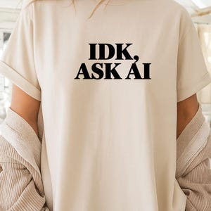 Könnte beinhalten: Ein beiges Kurzarm-T-Shirt mit dem schwarzen Text IDK, ASK AI in einer fetten Serifenschrift auf der Brust. Das Kleidungsstück hat einen Rundhalsausschnitt und umgekrempelte Ärmel und wird über einer beigen, gerippten Strickjacke getragen.