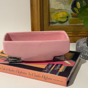 May include: A rectangular pink ceramic planter with a smooth finish, supported by two small, textured black and white striped feet. The planter is resting on a book titled Audrey: The Life of Audrey Hepburn by Charles Higham.