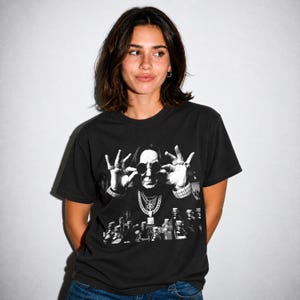 May include: A black short-sleeve t-shirt featuring a monochromatic graphic print of a person wearing round sunglasses, multiple layered necklaces, and rings, with several small glass bottles arranged in the foreground below the figure.