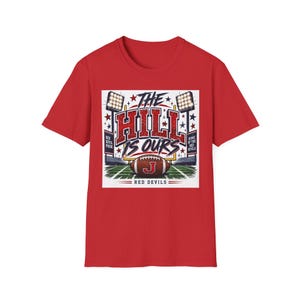 May include: A red short-sleeve t-shirt featuring a graphic print of a football field with goalposts, stadium lights, and a football with the letter J. The text reads THE HILL IS OURS, RED DEVIL PRIDE, HOME OF THE RED DEVILS, and RED DEVILS.