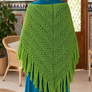 Puede incluir: Un chal triangular de ganchillo en un vibrante color verde lima, con un intrincado patrón calado con una columna central vertical y largos flecos colgantes a lo largo de los dos bordes inferiores, sobre una prenda de color azul cerceta.