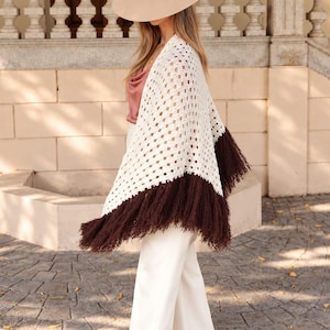 Puede incluir: Un chal de ganchillo color crema con un diseño de punto de patrón cuadrado y un borde ancho de flecos de color marrón oscuro. El chal se lleva sobre un top de satén malva y pantalones blancos de pierna ancha, con un sombrero de fieltro beige.