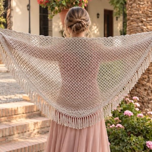 Puede incluir: Un chal triangular de ganchillo en color beige claro con un patrón calado y largos flecos en el borde inferior. El chal está drapeado sobre los hombros y los brazos, mostrando la intrincada textura del hilo y el detalle de los flecos anudados.