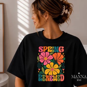 Könnte beinhalten: Ein schwarzes T-Shirt mit einem farbenfrohen Grafikdruck und dem Text SPRING RENEWED. Das Design zeigt große rosa, orange und gelbe Blumen mit grünen Blättern sowie den kleineren Text NEW SEASON BRIGHTER DAYS und SAME YOU GREATER THINGS.
