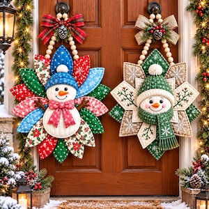 May include: Two decorative fabric snowman wall hangings featuring quilted petal-shaped backings. The left snowman wears a blue hat and red scarf, while the right snowman wears a green hat and scarf. Both are suspended by wooden bead loops.