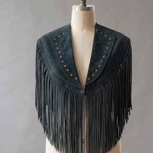 May include: A dark green suede capelet featuring long, flowing fringe along the hem and front edges. The garment is accented with a row of small, metallic studs lining the V-shaped neckline and front panels, creating a structured appearance.