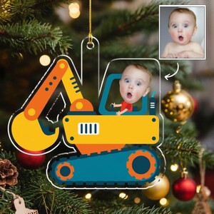 May include: A clear acrylic Christmas ornament shaped like a construction excavator. The excavator is yellow, orange, and blue, featuring a cutout window where a photograph of a baby with a surprised expression is displayed.