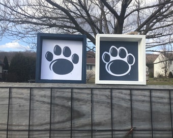May include: Two square wooden signs with a paw print design. The signs are painted in a dark blue and white color scheme. The paw print is a dark blue color on a white background on one sign and a white paw print on a dark blue background on the other sign.