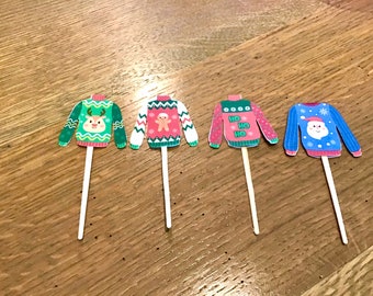 May include: Four Christmas sweater-themed cupcake toppers. Each topper features a different sweater design: a green sweater with a reindeer, a pink sweater with a gingerbread man, a pink sweater with "HO HO HO", and a blue sweater with Santa.