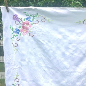 May include: White tablecloth with cross-stitched floral embroidery in pink, blue, and purple. The cloth is hanging on a clothesline with a wooden clothespin. The embroidery features detailed flower and leaf patterns.