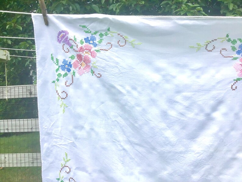 May include: White tablecloth with cross-stitched floral embroidery in pink, blue, and purple. The cloth is hanging on a clothesline with a wooden clothespin. The embroidery features detailed flower and leaf patterns.