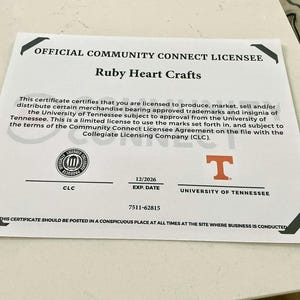 May include: A white certificate with black accents, featuring the text "OFFICIAL COMMUNITY CONNECT LICENSEE" and "Ruby Heart Crafts." Includes the University of Tennessee logo and a CLC seal. The expiration date is 12/2026.