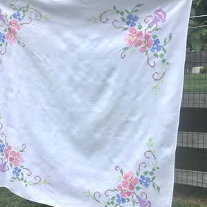 May include: White tablecloth with embroidered floral corner designs. The embroidery features pink, blue, and purple flowers with green leaves and stems. The tablecloth is hanging outside.