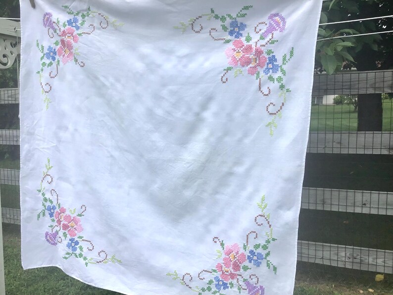 May include: White tablecloth with embroidered floral corner designs. The embroidery features pink, blue, and purple flowers with green leaves and stems. The tablecloth is hanging outside.