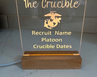 May include: A clear acrylic sign with the words "The Crucible" and the Marine Corps emblem. The sign also has the text "Recruit Name", "Platoon", and "Crucible Dates". The sign is lit from behind and sits on a wooden base.