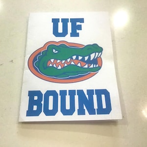 May include: A white paper with the words "UF BOUND" in blue block letters. The "UF" is above a green alligator logo with orange and blue accents. The alligator has white teeth and is set inside an orange oval.