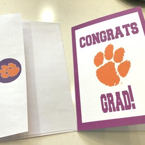 May include: A graduation card with a purple border and the words "CONGRATS GRAD!" in purple. An orange paw print is in the center. A matching envelope with a purple sticker featuring an orange paw print is also included.