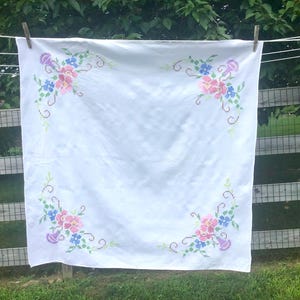 May include: White tablecloth with embroidered floral corner designs. The embroidery features pink, blue, and purple flowers with green vines. The tablecloth is hanging on a clothesline outdoors.