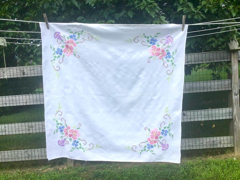 May include: White tablecloth with embroidered floral corner designs. The embroidery features pink, blue, and purple flowers with green vines. The tablecloth is hanging on a clothesline outdoors.