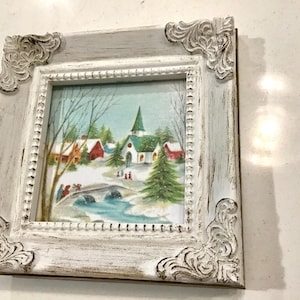 May include: A white, distressed, wooden frame with a winter scene of a small town with a church and a bridge. The scene is painted in shades of blue, green, red, and white.