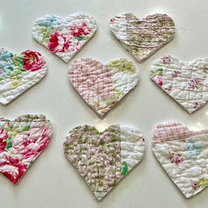 May include: Eight heart-shaped quilted fabric coasters with floral patterns in shades of pink, red, green, and blue. The coasters are arranged on a white surface, showcasing a variety of fabric designs.