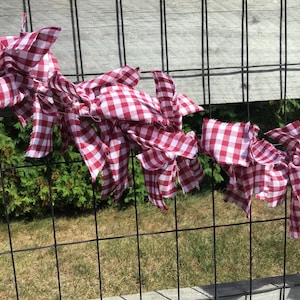 May include: A decorative garland made of red and white checkered fabric strips. The fabric is tied and draped across a black metal fence. The background includes green foliage and a wooden fence.