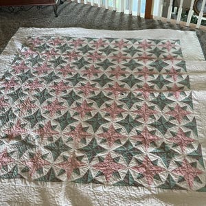 May include: A square quilt featuring a repeating star pattern in alternating shades of dusty pink and sage green. The design is set against a white background with a wide white border surrounding the central star-patterned section of the textile.
