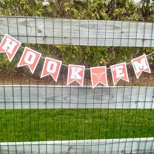 May include: A banner with orange and white pennants spelling "HOOK'EM" is strung across a wooden fence. The banner features the phrase in bold white letters with a longhorn symbol. The background includes green grass and foliage.