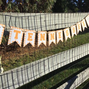 May include: A banner with the word "TENNESSEE" in orange letters on white pennants with an orange and white checkered border. The banner is strung across a wooden fence with a wire mesh backing. The first pennant has a dog illustration.