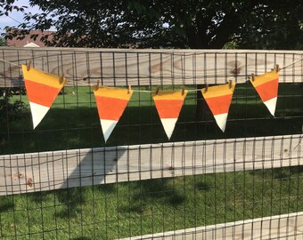 May include: A bunting banner featuring five triangular pennants hanging from a thin twine string. Each pennant is painted in horizontal stripes of yellow, orange, and white, mimicking the appearance of candy corn, and secured with small wooden clips.