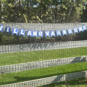 May include: A blue and white banner that reads "VILLANOVABOUND" is strung across a wooden fence. The banner is made of pennant-shaped flags with white letters on a blue background. The fence is made of weathered wood and wire mesh, with green grass in the background.