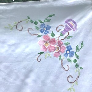 May include: White linen tablecloth with embroidered floral design. The embroidery features pink, blue, and purple flowers with green leaves and brown stems. The tablecloth is hanging from a clothesline.