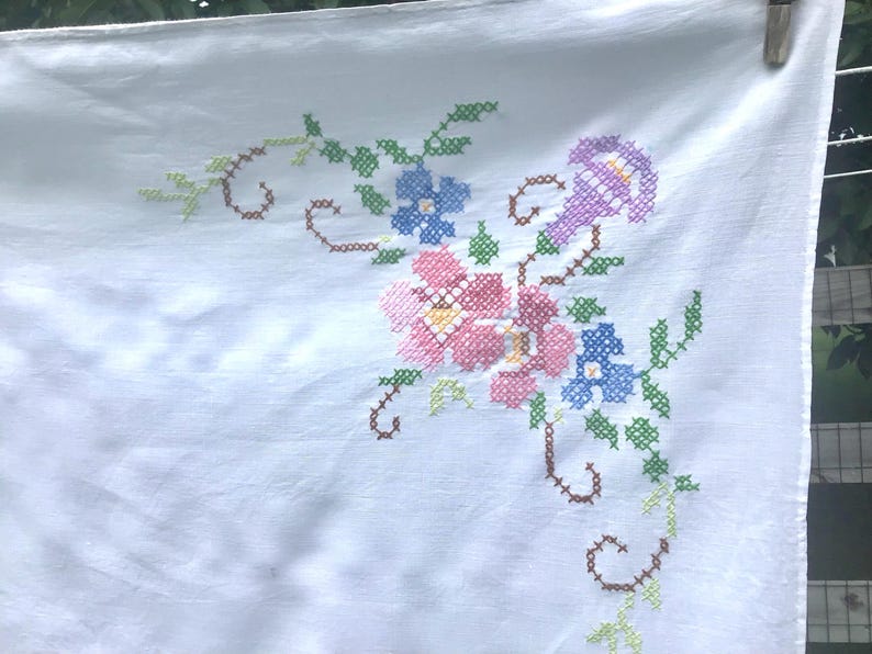 May include: White linen tablecloth with embroidered floral design. The embroidery features pink, blue, and purple flowers with green leaves and brown stems. The tablecloth is hanging from a clothesline.