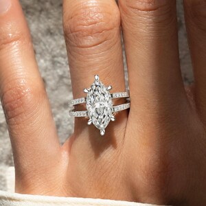 Peut inclure: Une bague argentée dotée d'une grande pierre centrale taille marquise maintenue par six griffes. L'anneau est divisé en deux rangées parallèles, toutes deux incrustées de petites pierres claires serties pavé sur le pourtour.