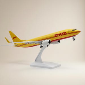Puede incluir: Un modelo de avión de carga DHL en amarillo y rojo montado sobre un soporte blanco. La aeronave presenta el logotipo de DHL en el fuselaje y la cola, con dos motores a reacción bajo las alas, tren de aterrizaje extendido y diseño aerodinámico.