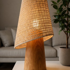 Pode incluir: Um candeeiro de mesa com uma base cilíndrica de madeira lisa com padrão de grão visível e um abajur cónico alto feito de material de fibra natural entrançada de cor clara que emite um brilho quente e suave da fonte de luz interna.