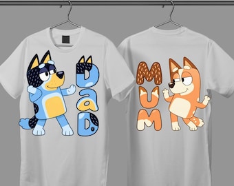 May include: Two white short-sleeve t-shirts hanging on hangers. The left shirt features a blue cartoon dog and the text DAD. The right shirt features an orange cartoon dog and the text MUM. Both shirts have a crew neckline and a straight hem.