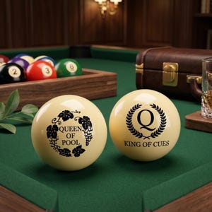 May include: Two cream-colored billiard balls featuring black printed text and graphics. One ball displays the text "QUEEN OF POOL" inside a floral wreath, while the other features a large "Q" inside a laurel wreath with "KING OF CUES" below.