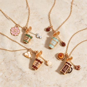 Peut inclure: Quatre colliers dorés avec des breloques miniatures sur le thème des boissons. L'un a une fleur rose et une tasse verte marquée Matcha Girlie, un autre une tasse vert clair marquée Mocha to Go, un autre une tasse marron marquée Coffee to Go et un mug marron marqué Brew.